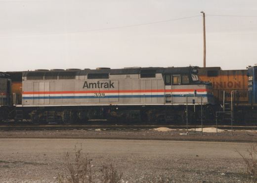 A silver Amtrak diesel locomotive with red, white and blue stripes and number 309 stands sideways in a rail yard, with a yellow Union Pacific locomotive and a blue locomotive behind it, under an overcast sky.