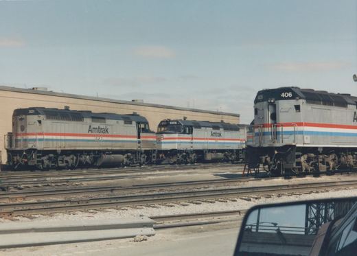 Three silver Amtrak diesel locomotives with red, white and blue stripes stand on yard tracks in front of a long tan building, and a car's side mirror shows in the lower right corner.