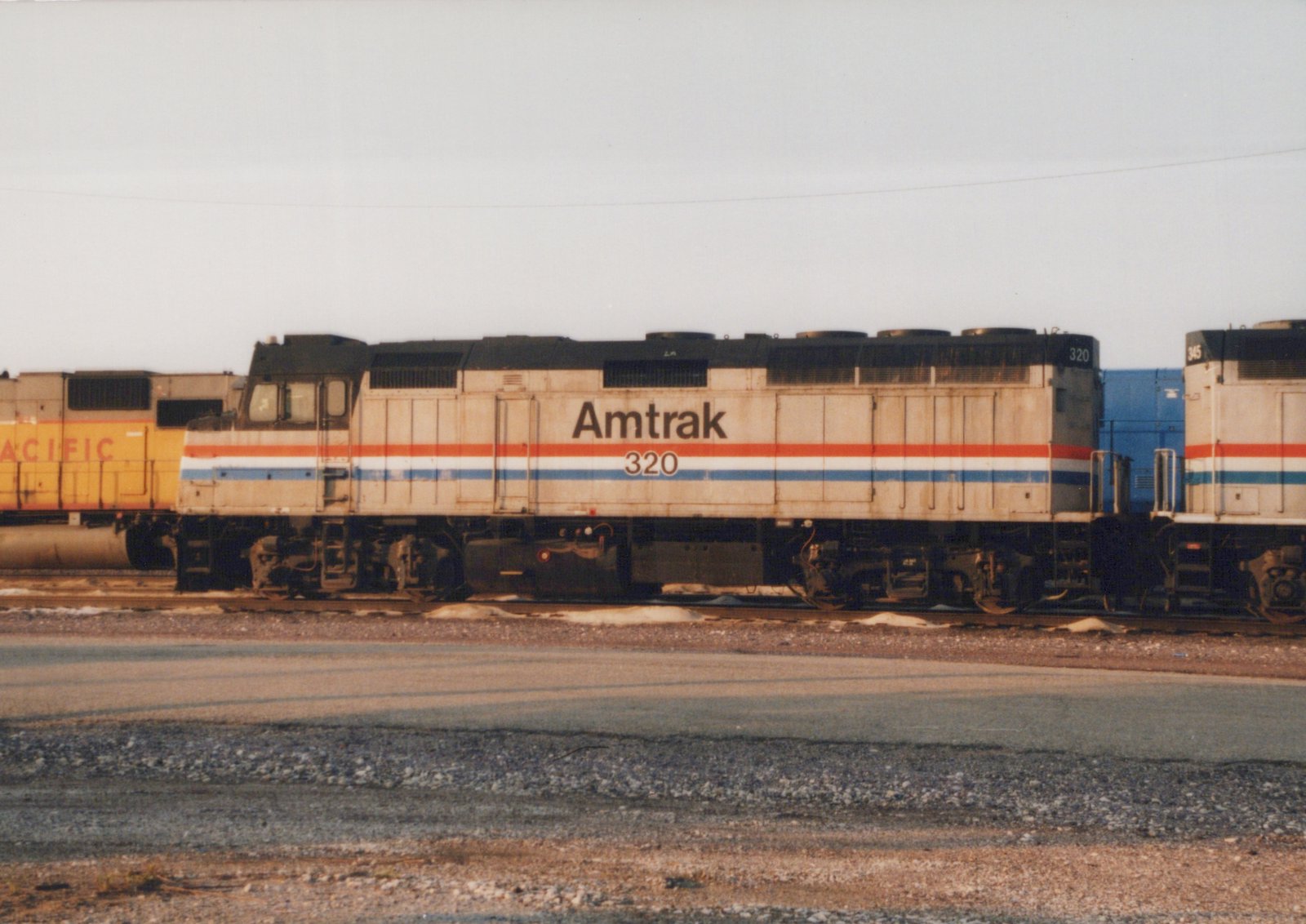 Side view of a silver Amtrak diesel locomotive numbered 320 with red, white and blue stripes, parked on a yard track with patches of snow, a yellow locomotive at left and another Amtrak unit at right.