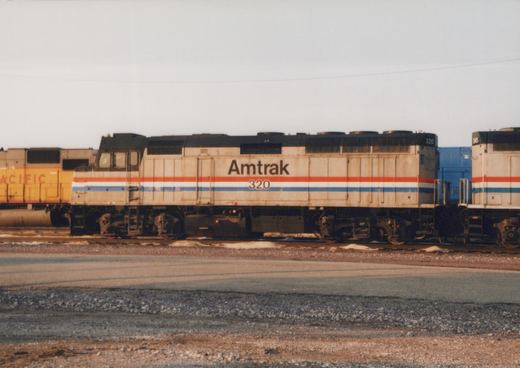 Side view of a silver Amtrak diesel locomotive numbered 320 with red, white and blue stripes, parked on a yard track with patches of snow, a yellow locomotive at left and another Amtrak unit at right.