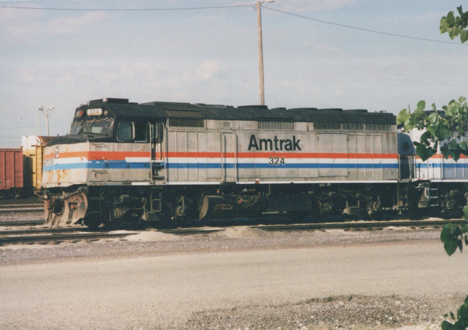 A side view of a weathered silver Amtrak diesel locomotive numbered 324, with red, white and blue stripes, sitting on a yard track with another striped Amtrak unit behind it and freight cars at the left.