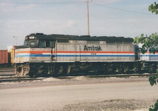 A side view of a weathered silver Amtrak diesel locomotive numbered 324, with red, white and blue stripes, sitting on a yard track with another striped Amtrak unit behind it and freight cars at the left.