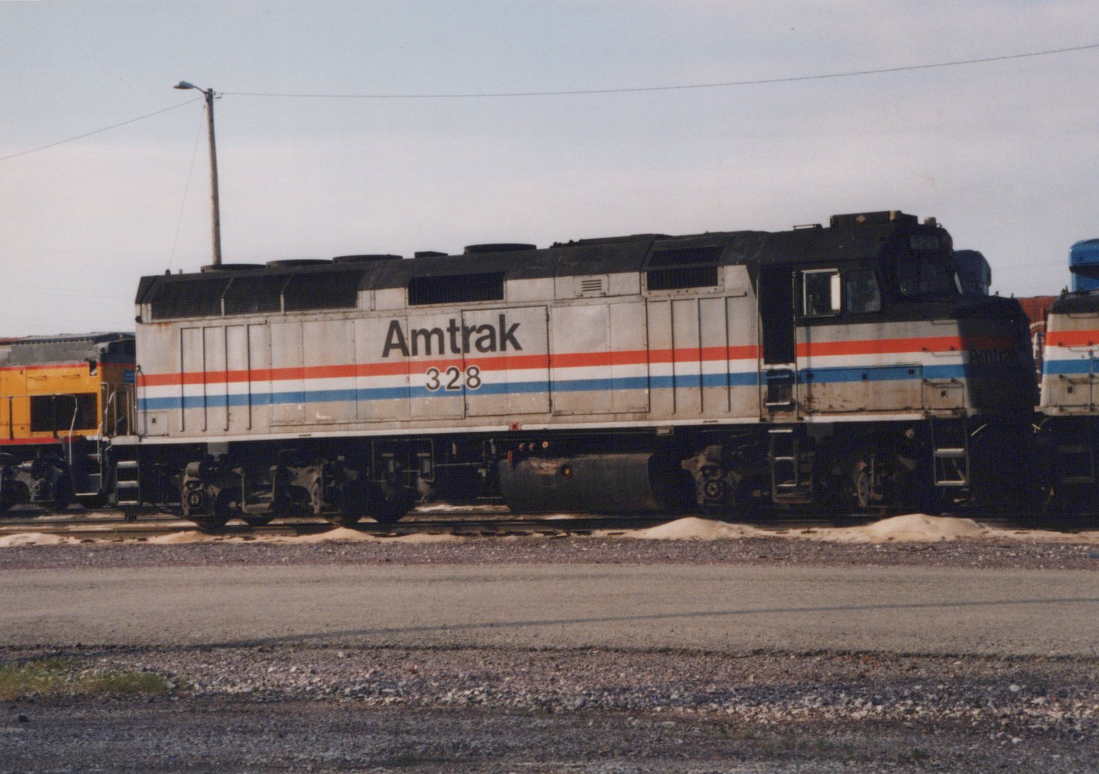Side view of a weathered silver Amtrak diesel locomotive numbered 328, with red, white and blue stripes. A yellow locomotive is to its left and another Amtrak unit is to its right, beside a gravel road.