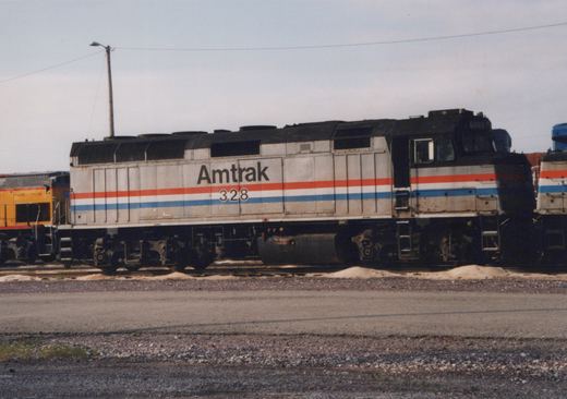 Side view of a weathered silver Amtrak diesel locomotive numbered 328, with red, white and blue stripes. A yellow locomotive is to its left and another Amtrak unit is to its right, beside a gravel road.