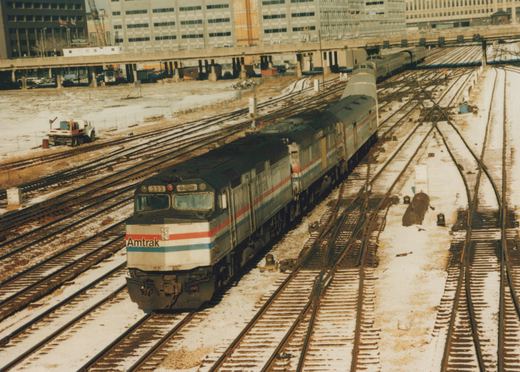 A silver Amtrak diesel locomotive numbered 333, with red, white and blue stripes, leads a second locomotive, a baggage car and passenger cars along snow-dusted tracks, with an elevated roadway and large buildings behind.