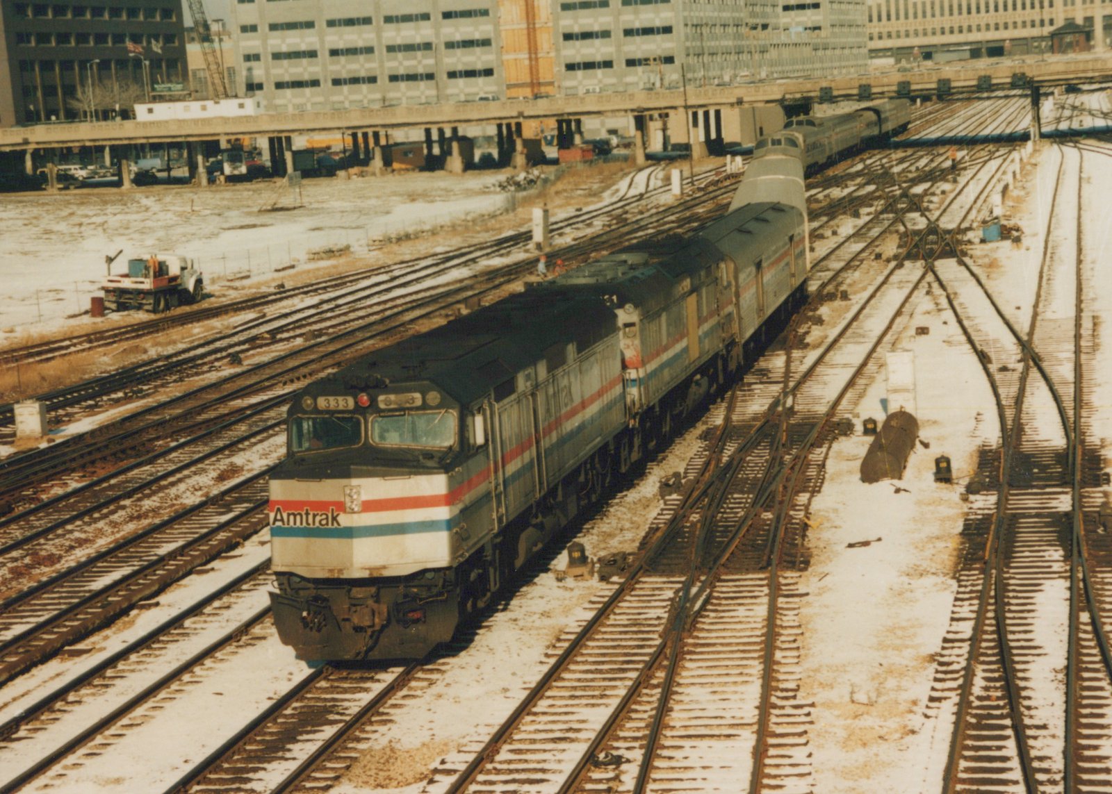 A silver Amtrak diesel locomotive numbered 333, with red, white and blue stripes, leads a second locomotive, a baggage car and passenger cars along snow-dusted tracks, with an elevated roadway and large buildings behind.
