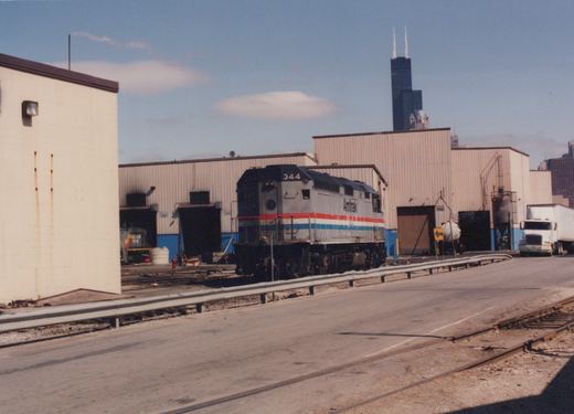 A silver locomotive with red, white and blue stripes and the number 344 stands in front of tan corrugated metal shop buildings. A tall black skyscraper with two antennas rises behind them under a blue sky, and a road with a guardrail and embedded rails runs across the foreground.