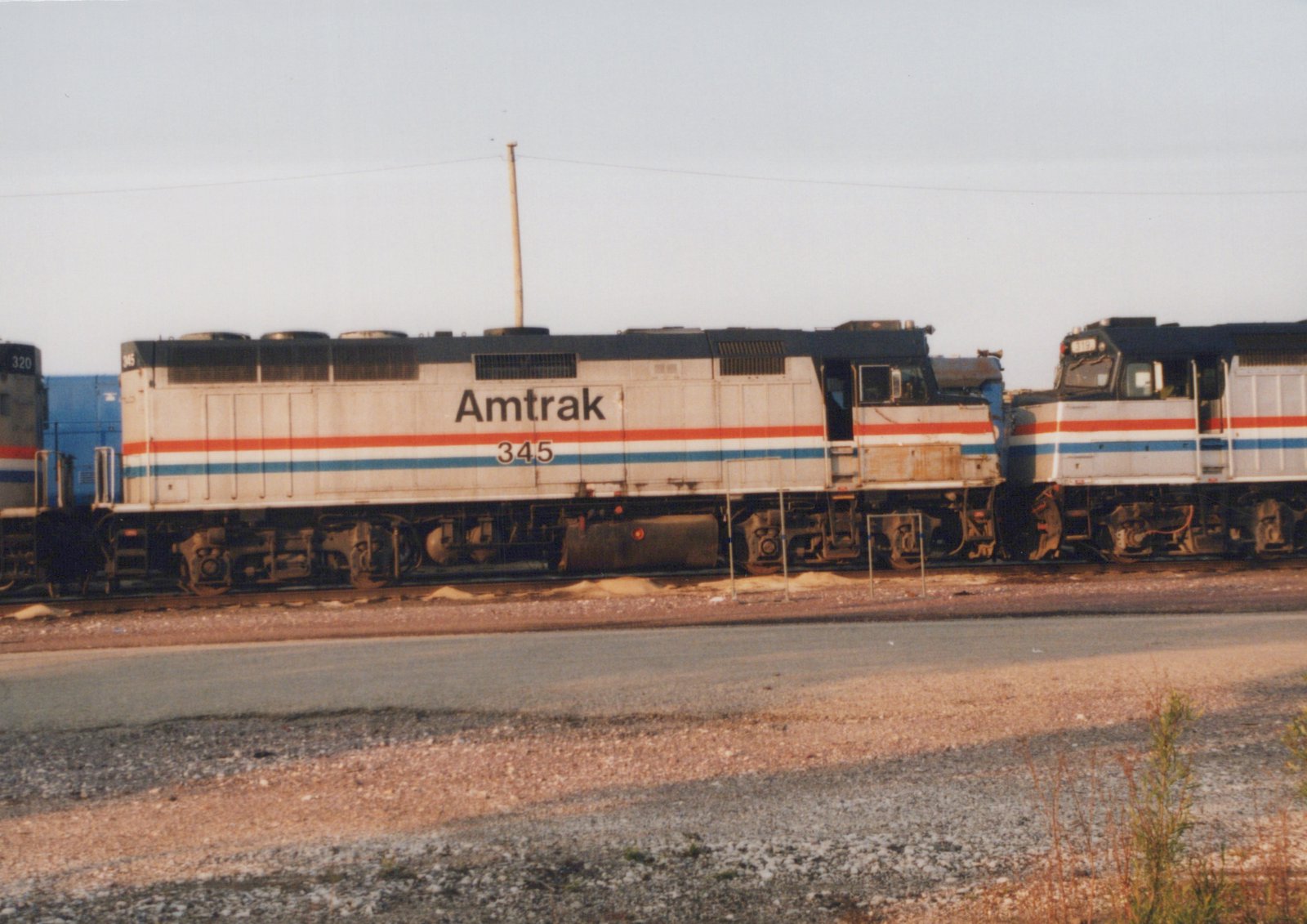 Side view of a gray locomotive with red, white and blue stripes lettered Amtrak 345, coupled between two similar locomotives on a yard track, with a gravel road in the foreground.