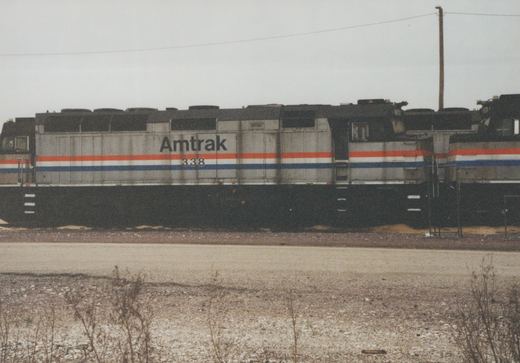 Side view of a grimy silver Amtrak diesel locomotive numbered 338 with red, white and blue stripes, coupled between two similar units beyond a gravel road under an overcast sky.