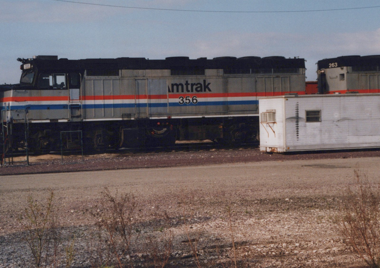 A silver diesel locomotive with a black roof and red, white and blue stripes, lettered Amtrak and numbered 356, stands in a yard beside a white trailer body, with a second locomotive numbered 263 behind it.