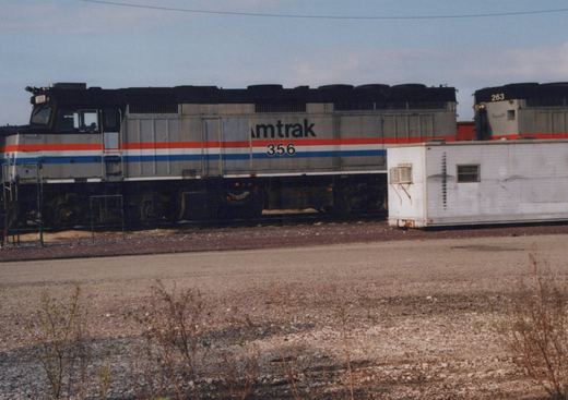 A silver diesel locomotive with a black roof and red, white and blue stripes, lettered Amtrak and numbered 356, stands in a yard beside a white trailer body, with a second locomotive numbered 263 behind it.