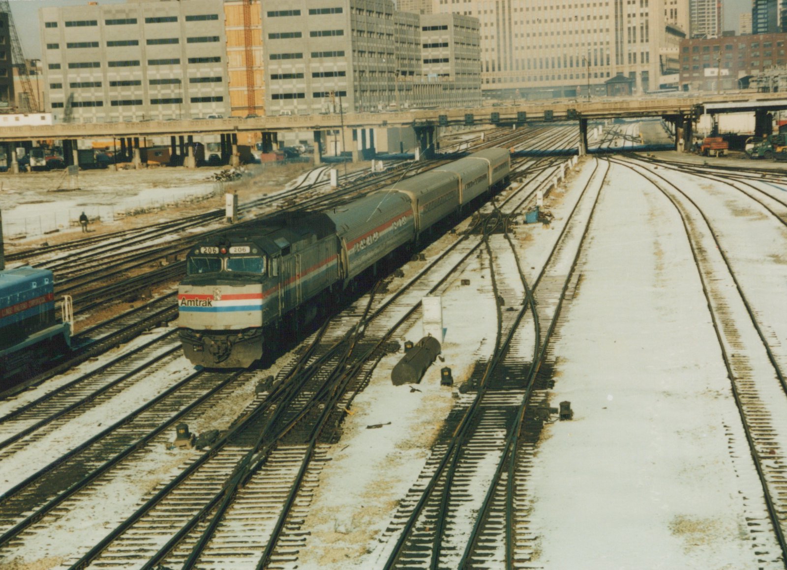 A silver Amtrak diesel locomotive with red, white and blue stripes pulls four silver passenger cars over a snowy maze of tracks and switches, with a street viaduct and tall office buildings behind.
