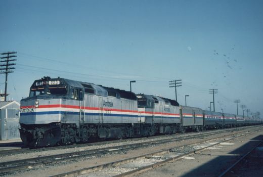 Two silver Amtrak diesel locomotives with red, white and blue stripes lead a long passenger train on one of several tracks under a clear blue sky, with a pale moon and a pole line overhead.