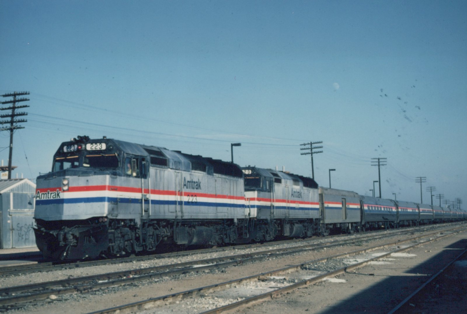 Two silver Amtrak diesel locomotives with red, white and blue stripes lead a long passenger train on one of several tracks under a clear blue sky, with a pale moon and a pole line overhead.