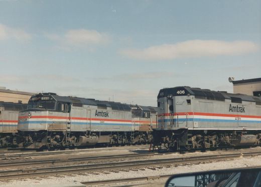 Silver Amtrak diesel locomotives with red, white and blue stripes are parked on yard tracks under a partly cloudy sky. A car's side mirror shows in the lower right corner.