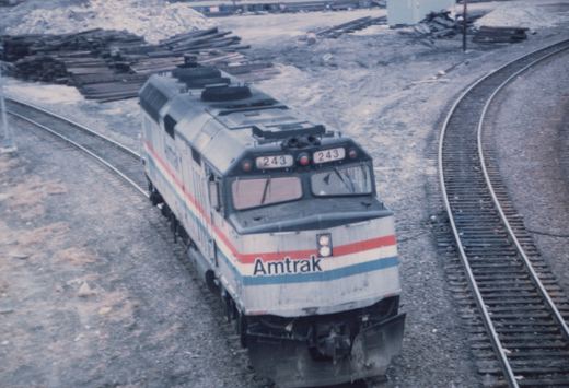 Overhead view of a silver Amtrak diesel locomotive numbered 243, with red, white and blue stripes, on a curving track next to a second track and piles of railroad ties.