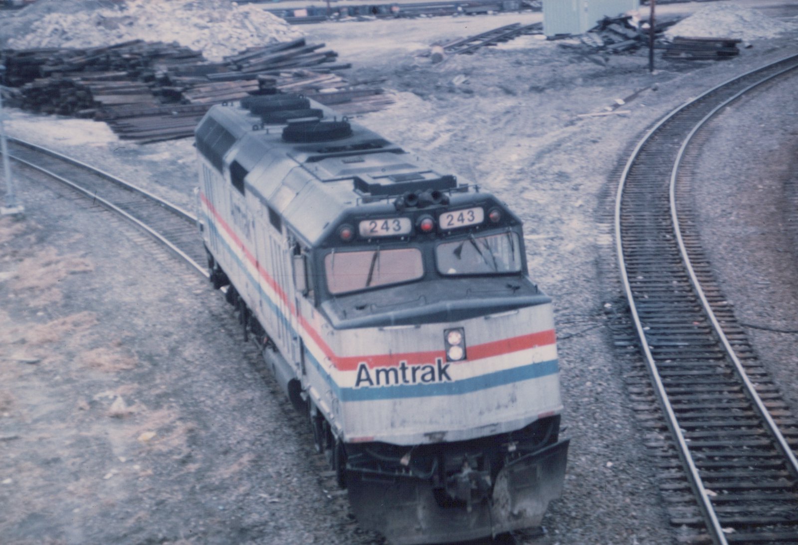 Overhead view of a silver Amtrak diesel locomotive numbered 243, with red, white and blue stripes, on a curving track next to a second track and piles of railroad ties.