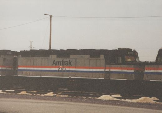 Side view of a silver diesel locomotive lettered Amtrak 263, with red, white and blue stripes, coupled between two similar locomotives on a yard track under an overcast sky.