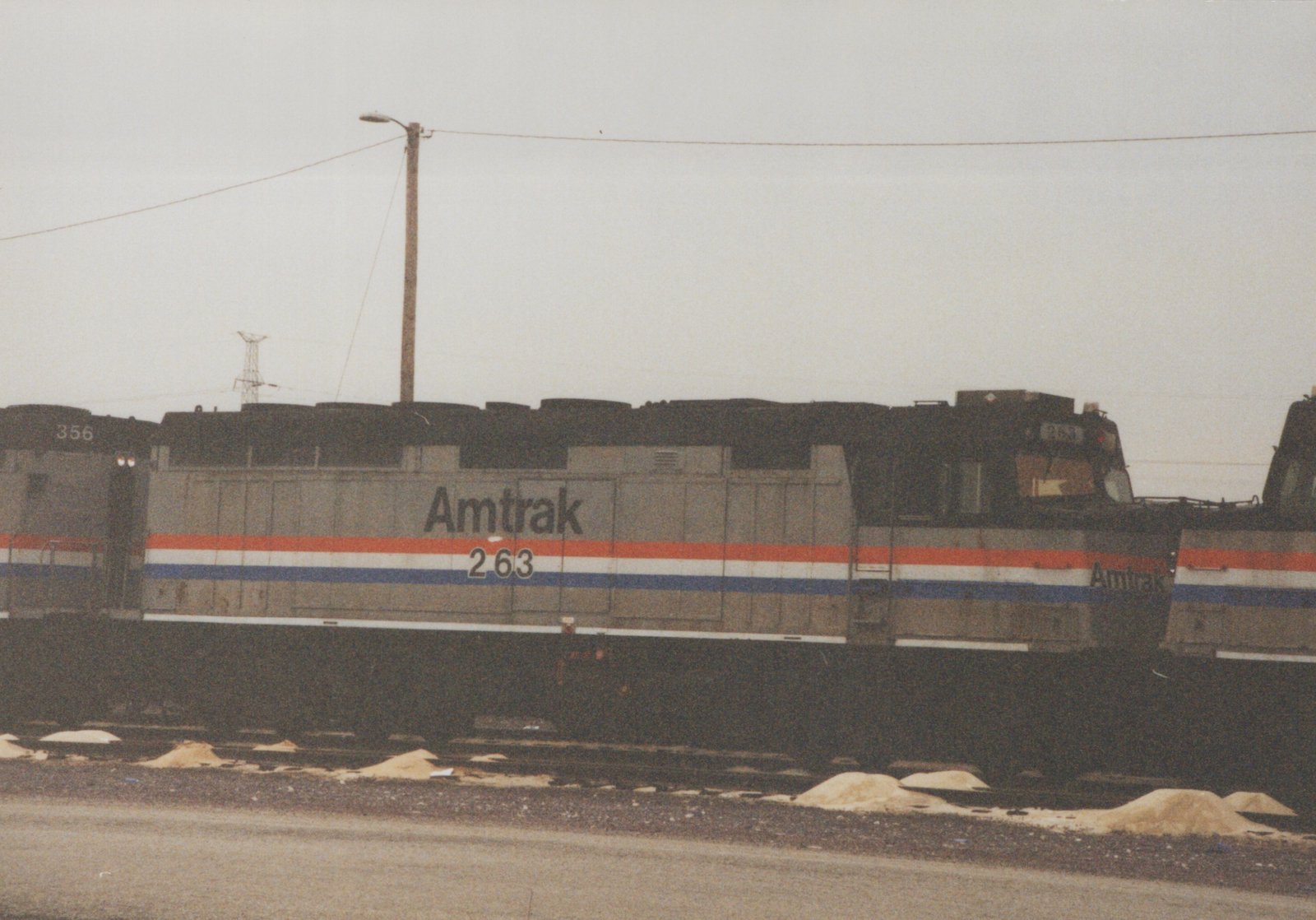 Side view of a silver diesel locomotive lettered Amtrak 263, with red, white and blue stripes, coupled between two similar locomotives on a yard track under an overcast sky.