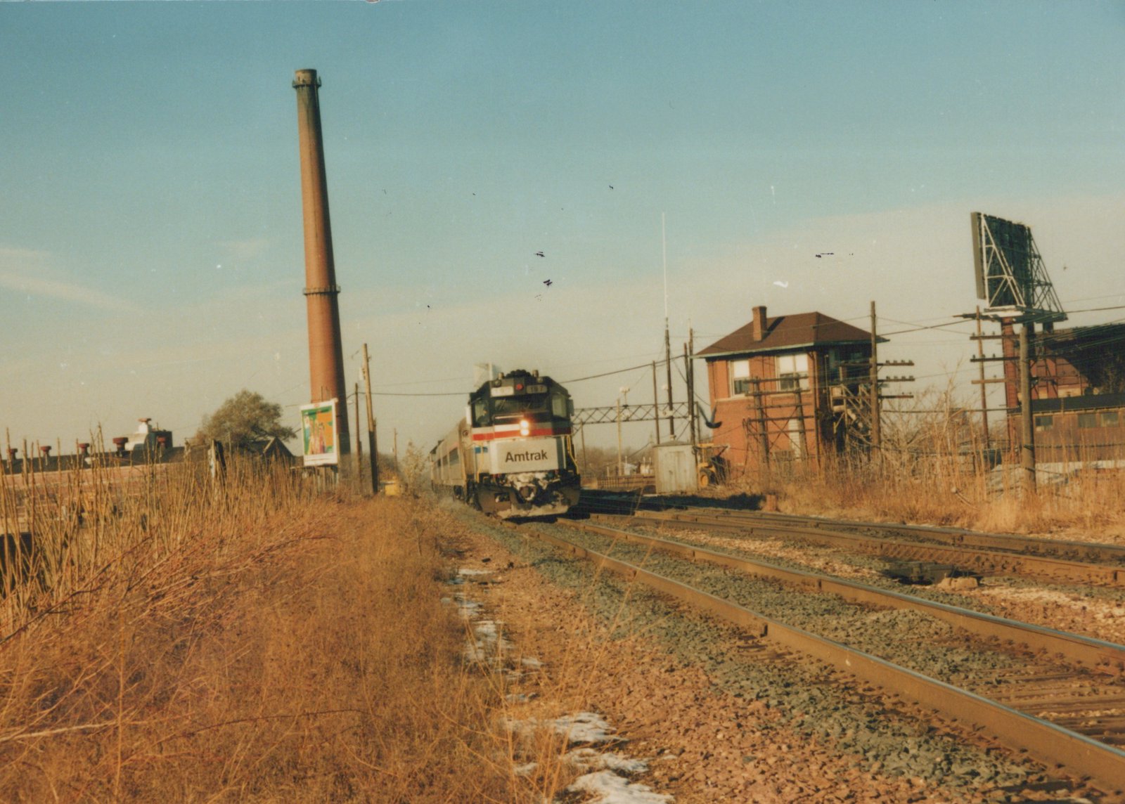 A silver Amtrak diesel locomotive with a red, white and blue stripe leads stainless steel passenger cars along double track, past a brick interlocking tower, a signal bridge and a tall brick smokestack, with dry winter grass and patches of snow in the foreground.