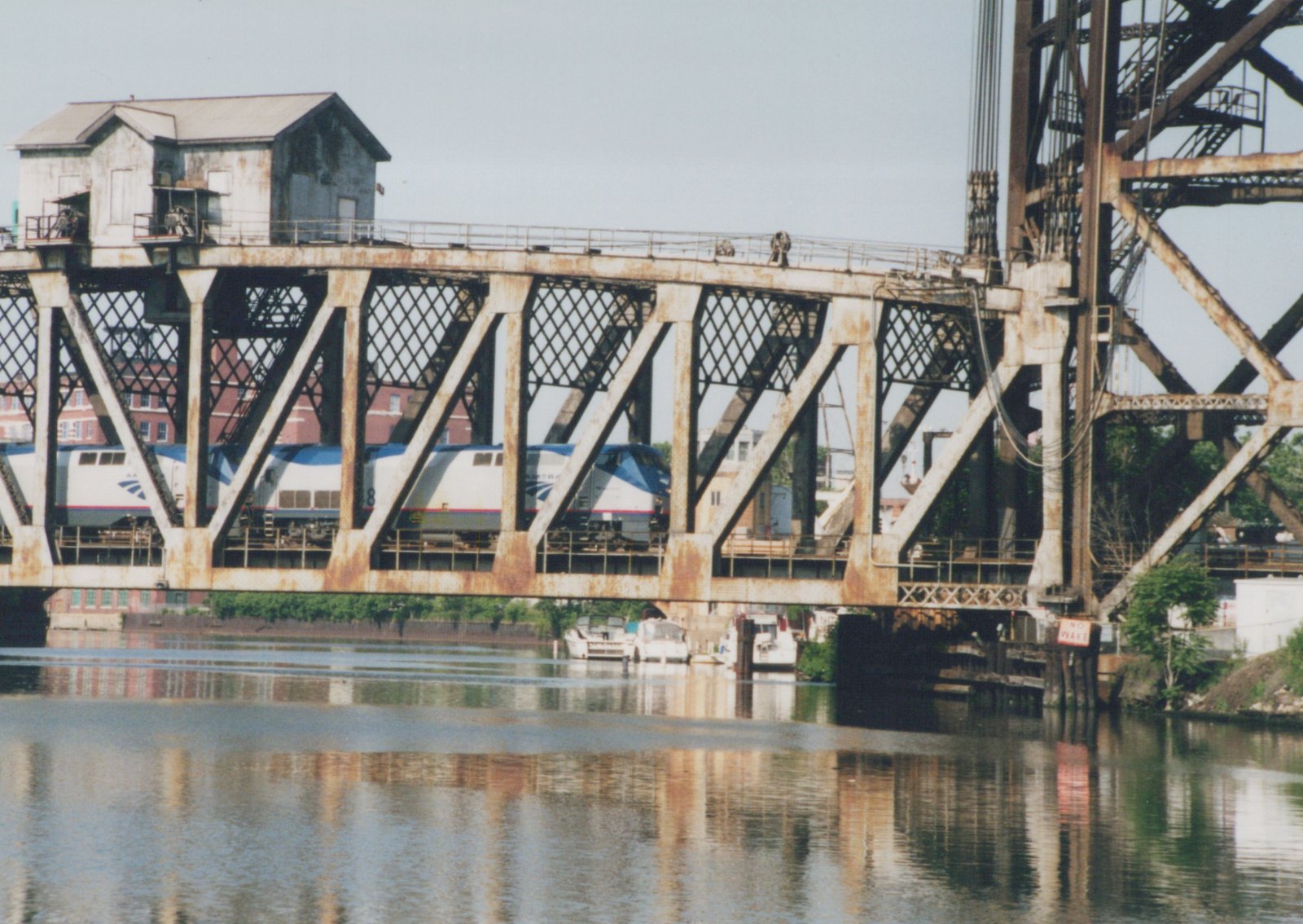 Two silver and blue Amtrak locomotives seen through the diagonal members of a rusty steel truss bridge with a small operator's house on top, a lift tower at right and calm water reflecting the bridge below.