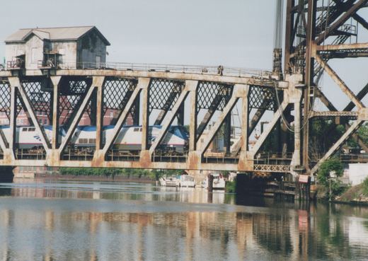 Two silver and blue Amtrak locomotives seen through the diagonal members of a rusty steel truss bridge with a small operator's house on top, a lift tower at right and calm water reflecting the bridge below.