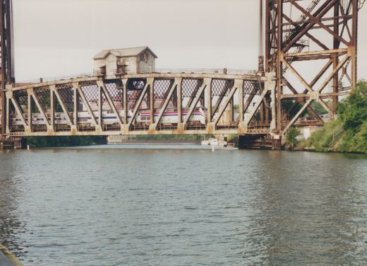 A silver passenger train with red, white and blue stripes crosses a rusty steel truss bridge with a small operator's house on top, beside a tall steel lift tower above a wide river.