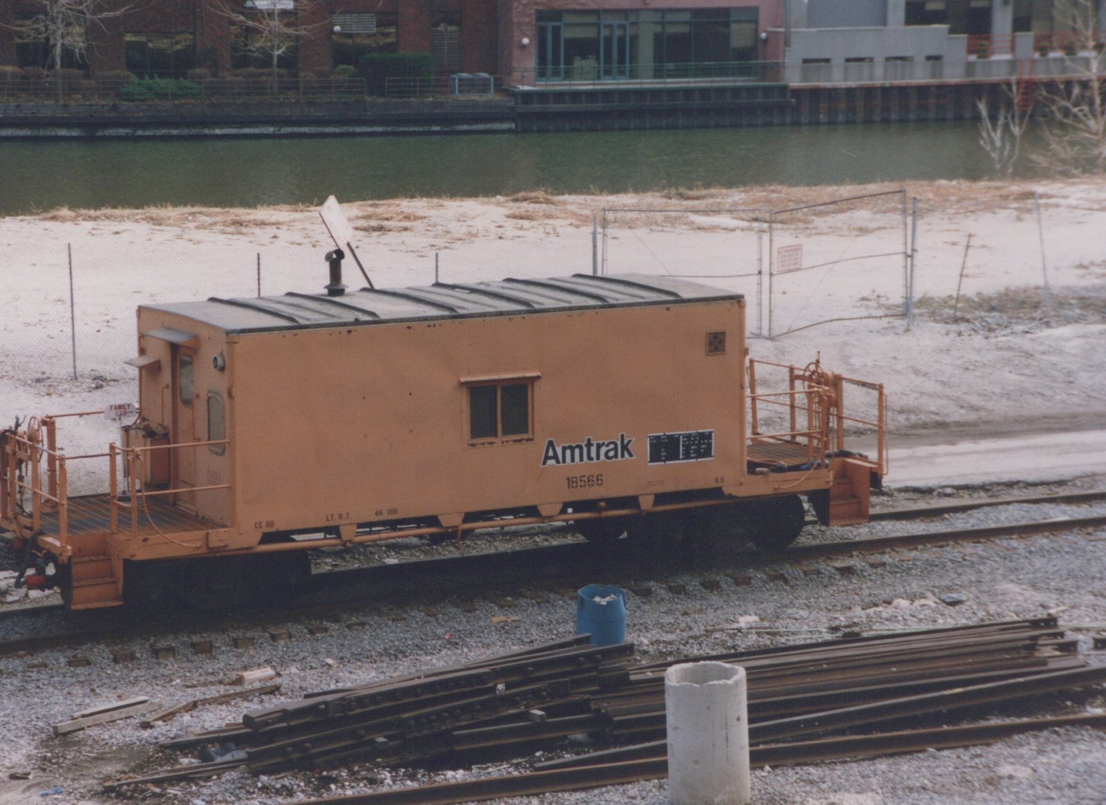 An orange caboose with a short box cabin, open railed end platforms and black Amtrak lettering stands on a track, with stacked rails in the foreground and a river and modern buildings behind.
