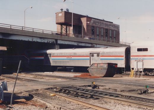 A silver Amtrak baggage car with red, white and blue stripes stands on a yard track under a street viaduct, with a stainless bilevel coach-dorm car at right, a steel dumpster in front and a brick building lettered Industrial Gases above.