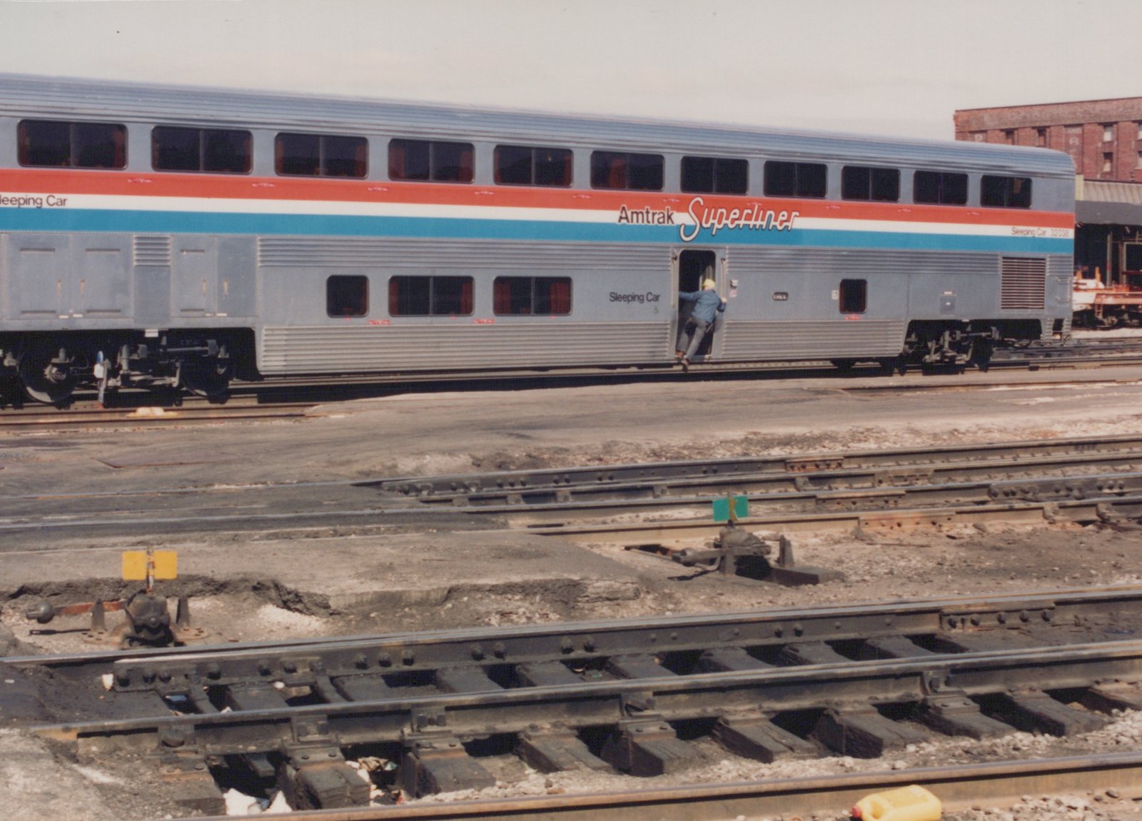 A stainless steel bi-level Amtrak sleeping car with red, white and blue stripes stands on a yard track while a worker climbs into its side door, with switch stands and track in the foreground.