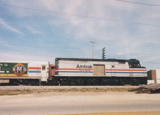 Side view of a silver Amtrak locomotive-bodied unit numbered 90368 with red and blue stripes and a roll-up side door, coupled to a coach with a green advertising wrap, on ballasted track beside a road under a partly cloudy sky.