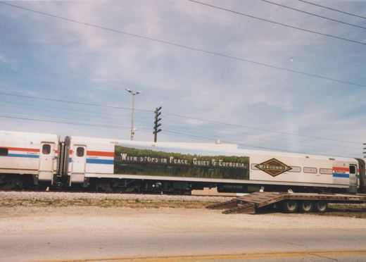 Side view of silver passenger cars with red, white and blue stripes. The middle car is wrapped with a green field scene, the slogan "With stops in Peace, Quiet & Euphoria" and a Wisconsin 1848-1998 logo. A lowboy trailer is parked beside the track and a road is in the foreground.