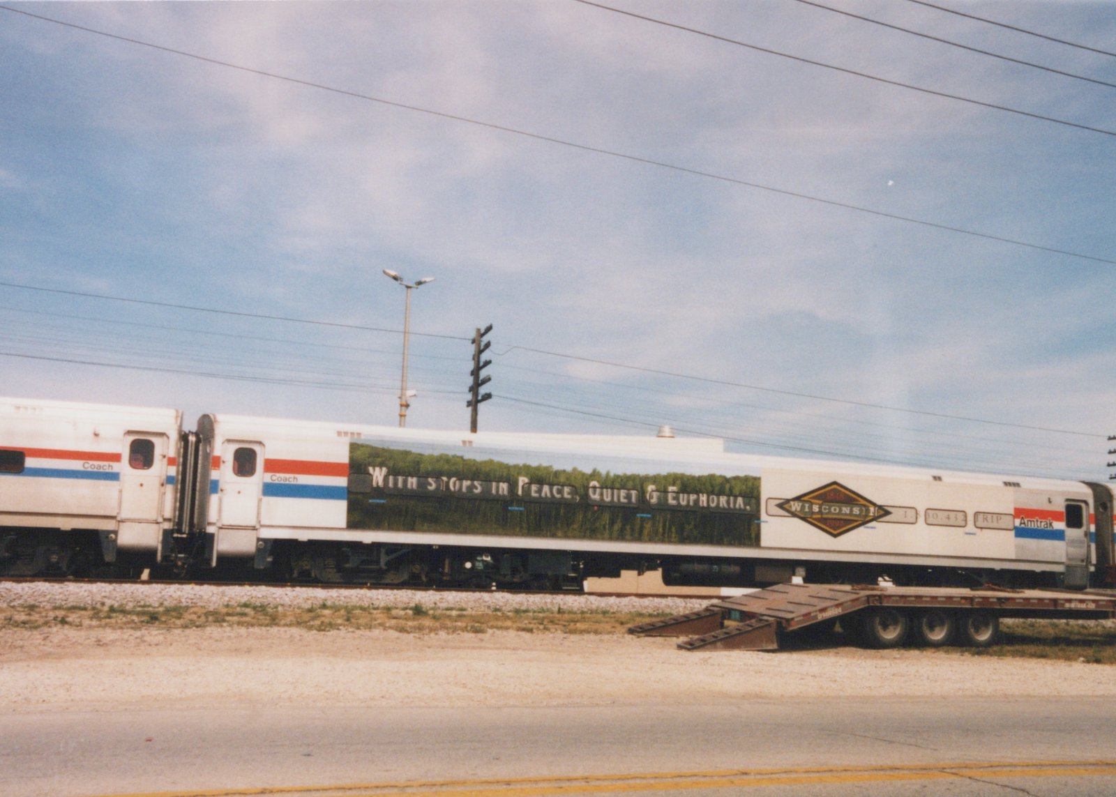 Side view of silver passenger cars with red, white and blue stripes. The middle car is wrapped with a green field scene, the slogan "With stops in Peace, Quiet & Euphoria" and a Wisconsin 1848-1998 logo. A lowboy trailer is parked beside the track and a road is in the foreground.
