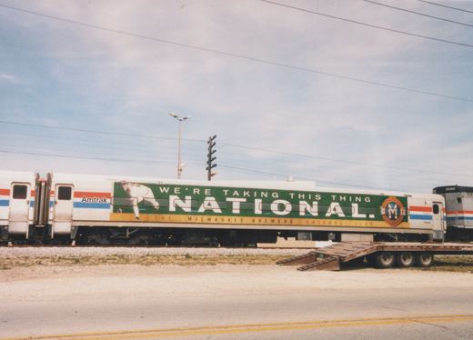 A stainless steel passenger coach with a green Milwaukee Brewers advertising wrap, coupled between another Amtrak coach and a silver unit numbered 90368, stands on a track beside a road with a lowboy trailer parked nearby.