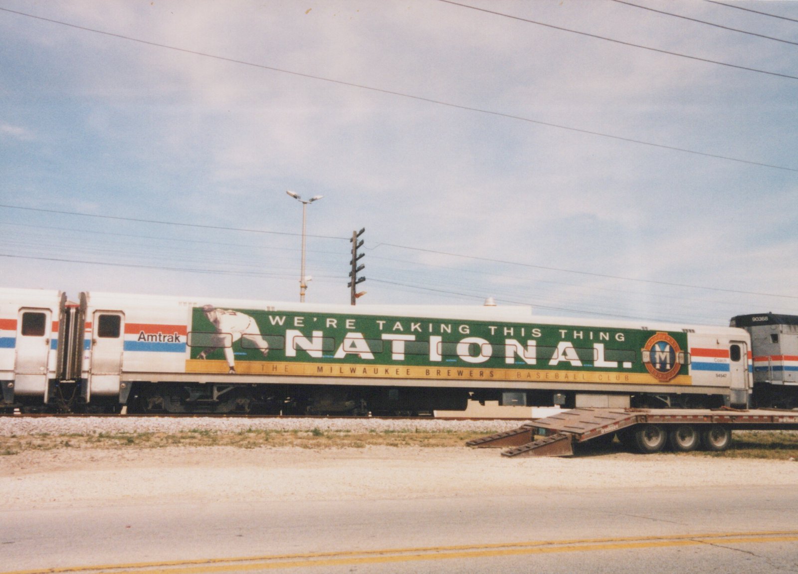 A stainless steel passenger coach with a green Milwaukee Brewers advertising wrap, coupled between another Amtrak coach and a silver unit numbered 90368, stands on a track beside a road with a lowboy trailer parked nearby.