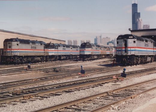 Several silver Amtrak diesel locomotives with red, white and blue stripes stand on yard tracks with blue STOP flags in the foreground and a tall black skyscraper in the background.