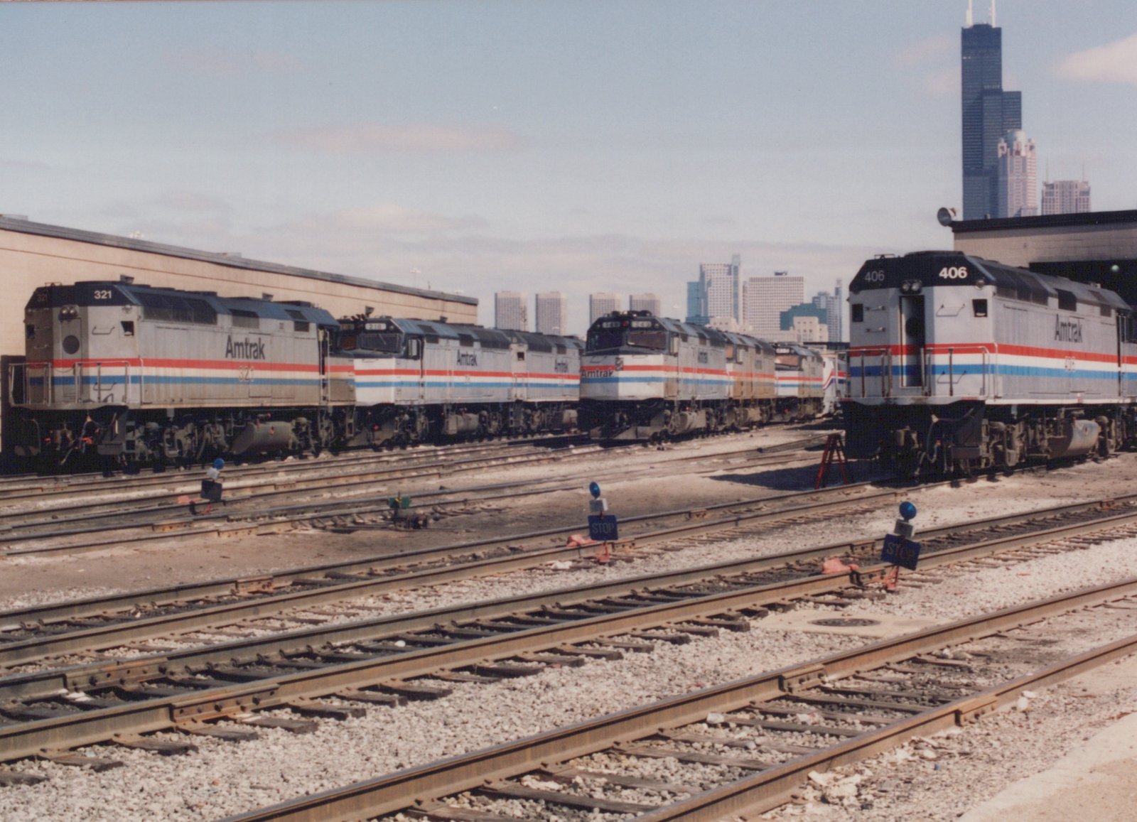 Several silver Amtrak diesel locomotives with red, white and blue stripes stand on yard tracks with blue STOP flags in the foreground and a tall black skyscraper in the background.