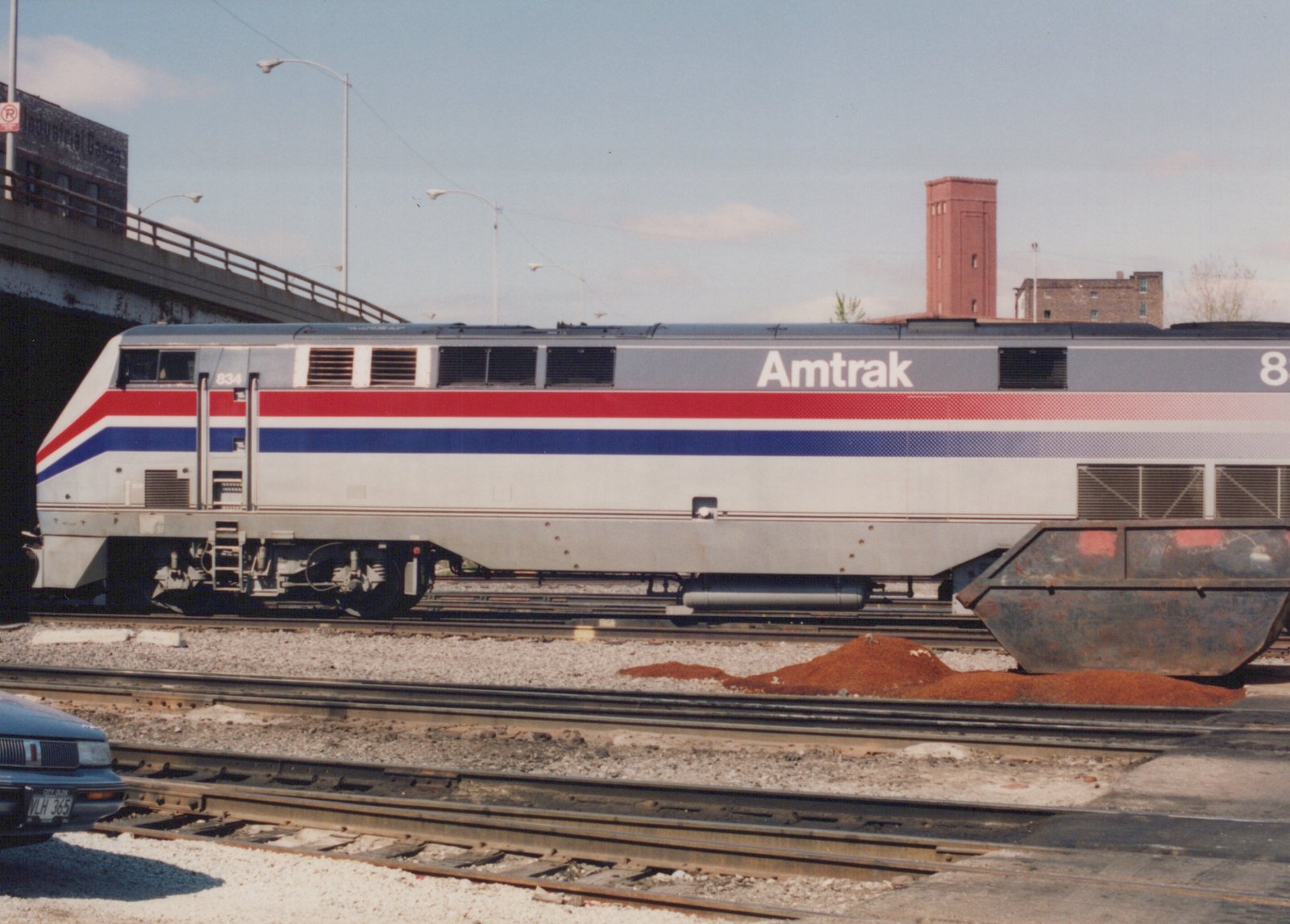 A silver diesel locomotive with red and blue stripes and Amtrak lettering stands in a rail yard below a street overpass, with a pile of red material and a rusty steel skip in front of it.