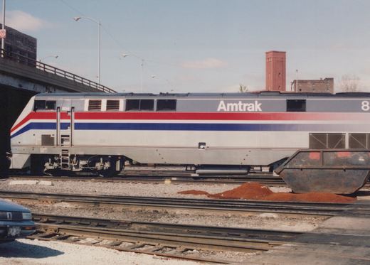 A silver diesel locomotive with red and blue stripes and Amtrak lettering stands in a rail yard below a street overpass, with a pile of red material and a rusty steel skip in front of it.