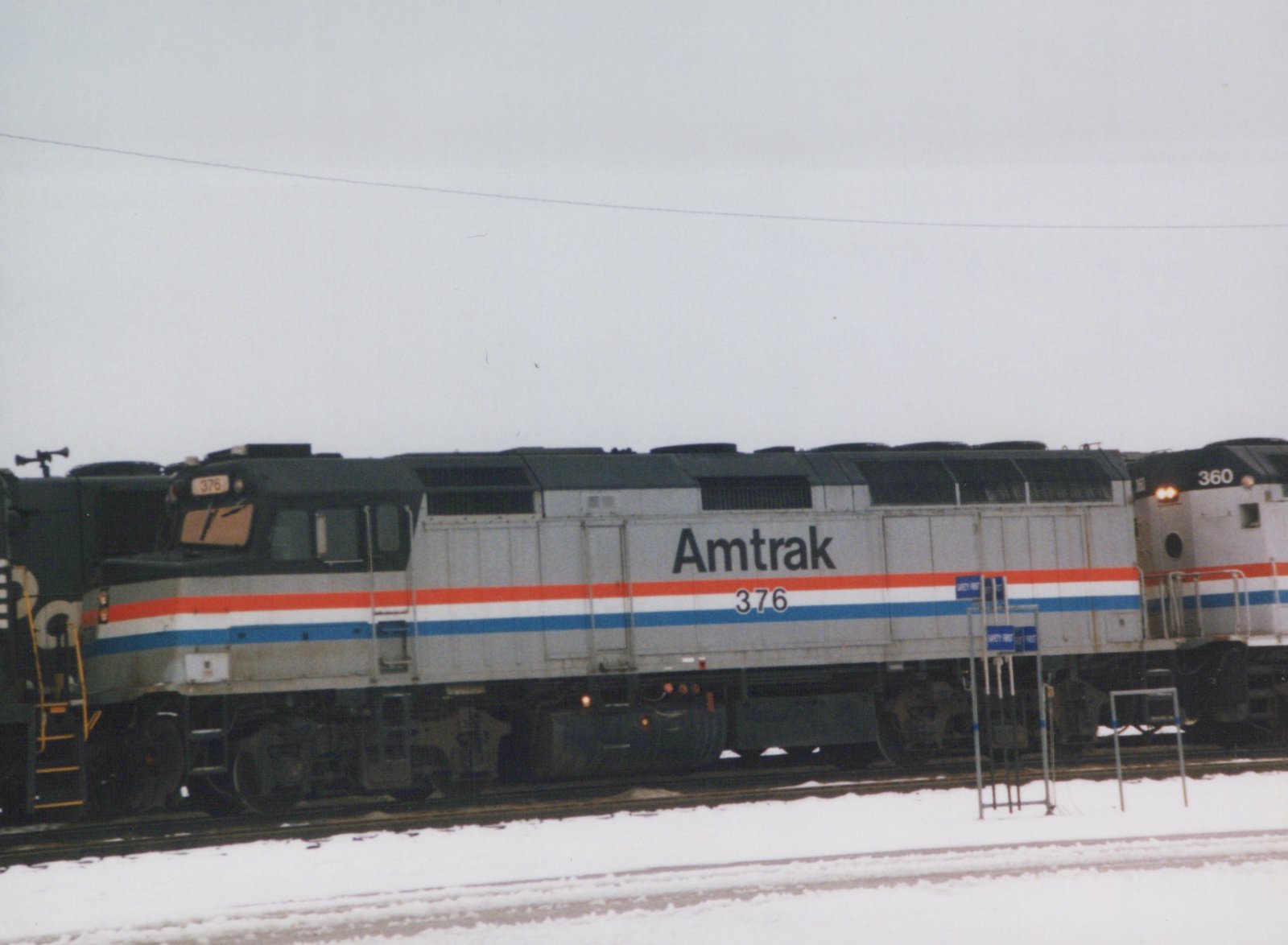 Side view of a silver diesel locomotive with red, white and blue stripes, lettered Amtrak 376, on snowy yard tracks under a gray sky, with another Amtrak unit numbered 360 at right and a dark green locomotive at left.
