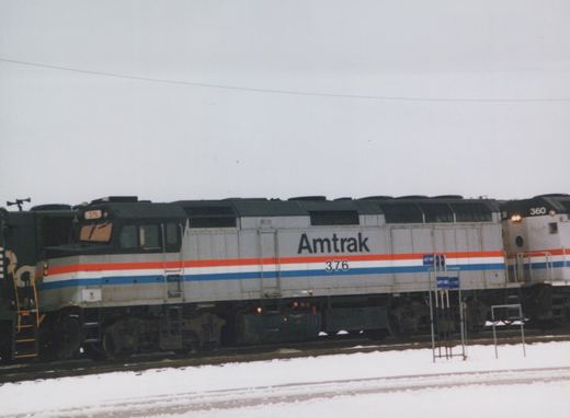 Side view of a silver diesel locomotive with red, white and blue stripes, lettered Amtrak 376, on snowy yard tracks under a gray sky, with another Amtrak unit numbered 360 at right and a dark green locomotive at left.