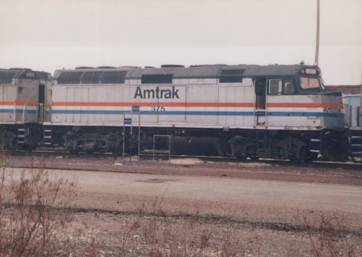 Side view of a silver diesel locomotive with red and blue stripes, lettered Amtrak and numbered 375, standing on a yard track with a second Amtrak unit, numbered 281, coupled behind it and dry weeds in the foreground.