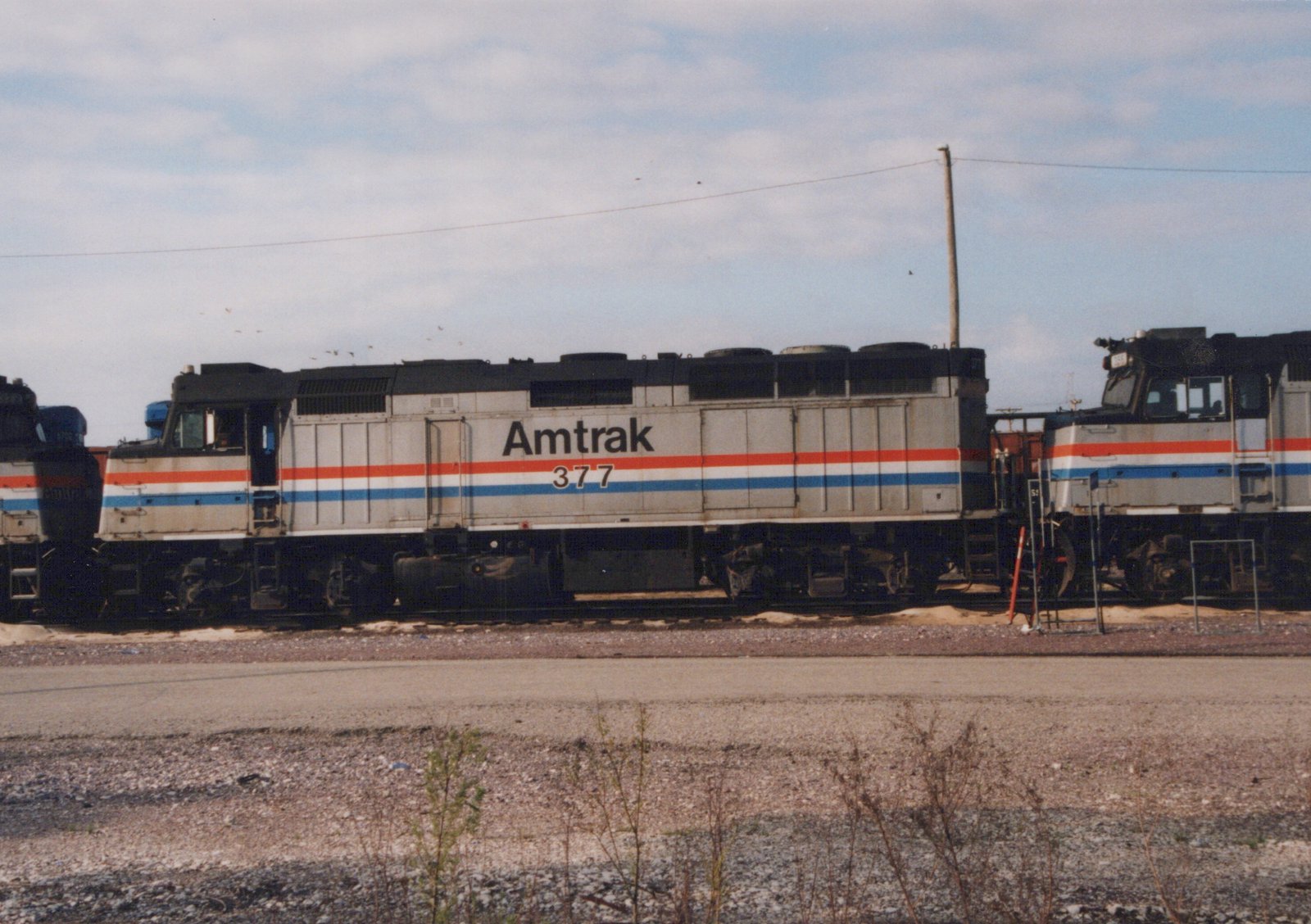 Side view of a silver Amtrak diesel locomotive numbered 377, with red, white and blue stripes, standing in a yard between two similar locomotives, with a gravel road and weeds in the foreground.