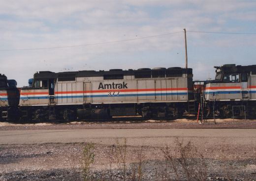 Side view of a silver Amtrak diesel locomotive numbered 377, with red, white and blue stripes, standing in a yard between two similar locomotives, with a gravel road and weeds in the foreground.