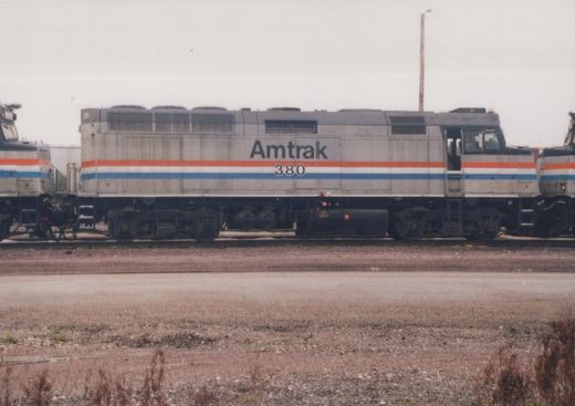 Side view of a silver diesel locomotive with red, white and blue stripes, lettered Amtrak 380, standing on a track between two similar locomotives under an overcast sky, with a gravel lot and weeds in front.