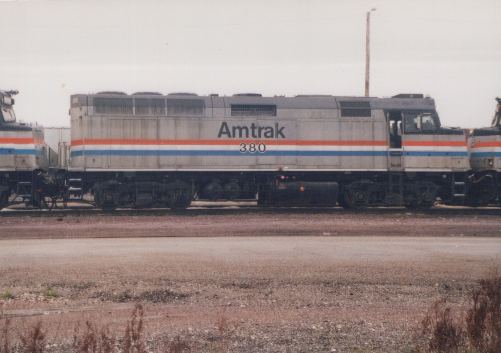 Side view of a silver diesel locomotive with red, white and blue stripes, lettered Amtrak 380, standing on a track between two similar locomotives under an overcast sky, with a gravel lot and weeds in front.