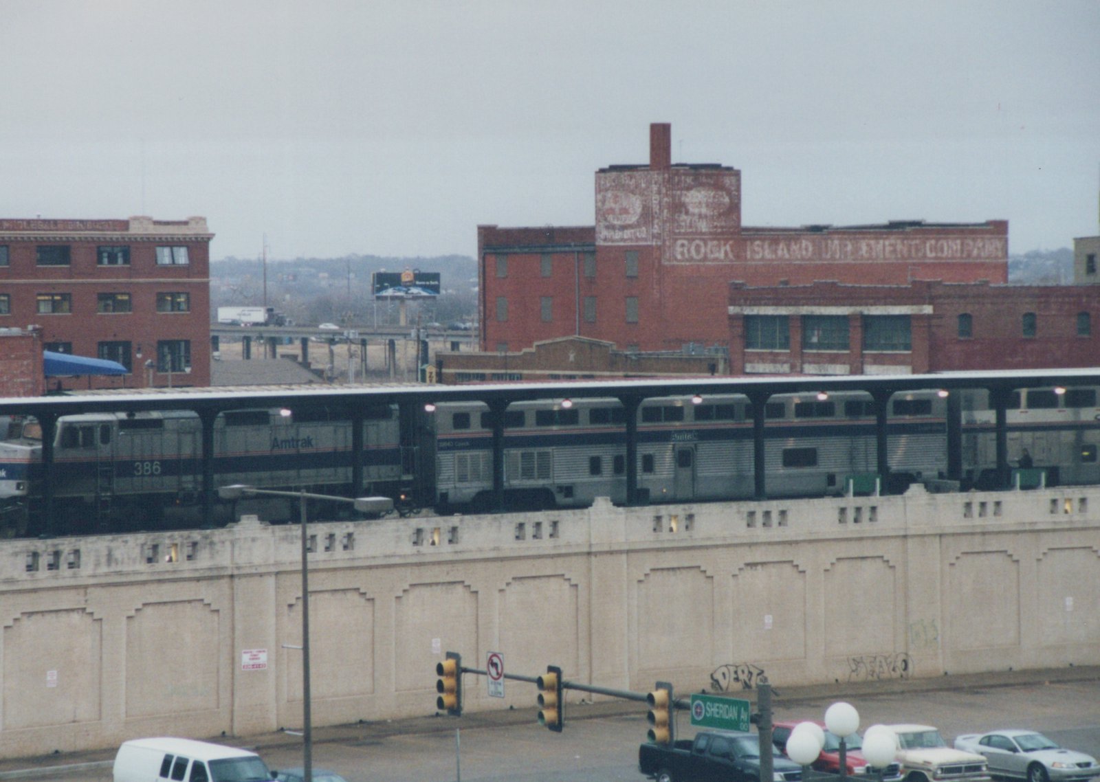 A silver Amtrak train with locomotive 386 and a two-level stainless steel coach under a long canopy on top of a concrete retaining wall, with old brick warehouses behind and traffic signals and parked pickups in the street below.
