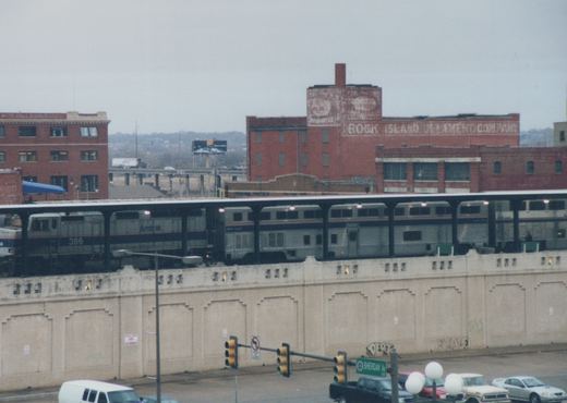 A silver Amtrak train with locomotive 386 and a two-level stainless steel coach under a long canopy on top of a concrete retaining wall, with old brick warehouses behind and traffic signals and parked pickups in the street below.