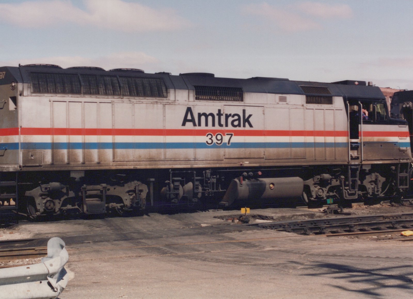 Side view of a silver diesel locomotive with red, white and blue stripes, lettered Amtrak and numbered 397, standing beside a paved crossing over yard tracks.