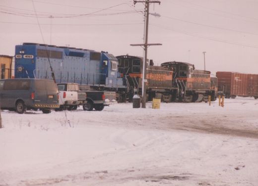 A blue diesel locomotive numbered 4002 and two black-and-orange switchers lettered HARBOR stand in a snow-covered yard, with a van and two pickup trucks parked in front and a red boxcar at right.